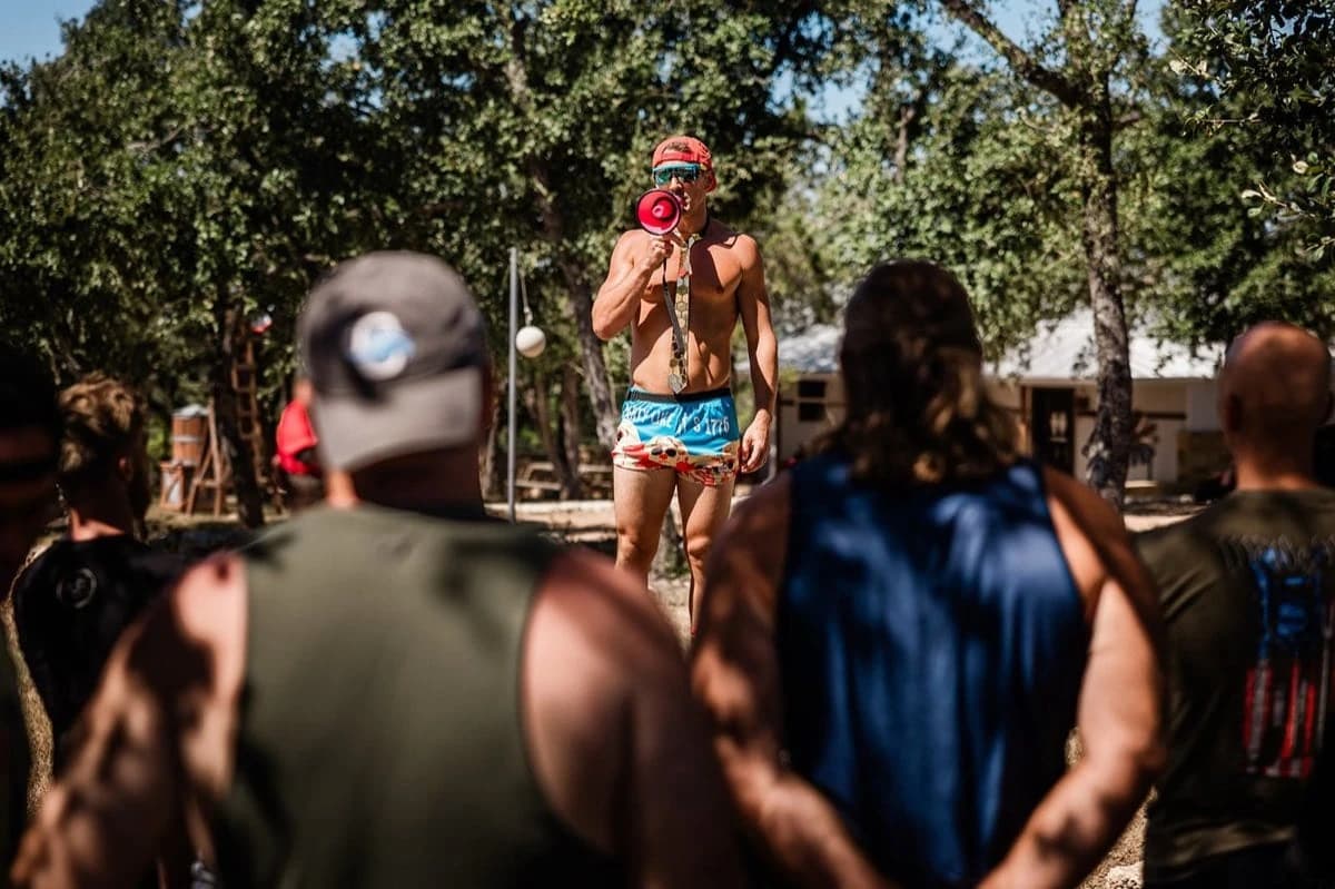 Jordan Bowditch in a cap and tie yelling through a megaphone at a crowd of men outdoors